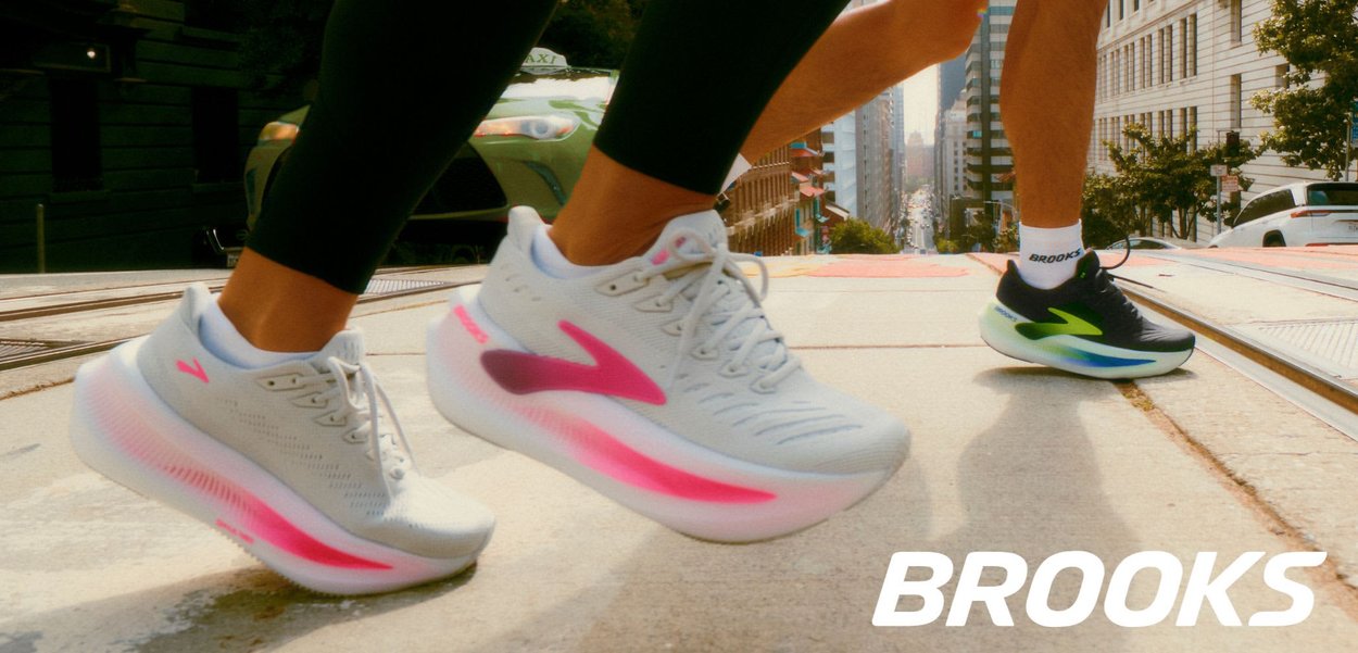 Scarpe da corsa Brooks con tomaia grigio chiaro, dettagli rosa e suole testurizzate. Corridori che attraversano una strada indossando leggings neri aderenti.