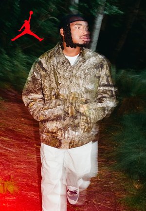 Tarnmuster-Jacke mit Reißverschluss und strukturiertem Rinden-Print, kombiniert mit weißen Hosen und dunklen Schuhen. Rotes Jumpman-Logo in der oberen linken Ecke.