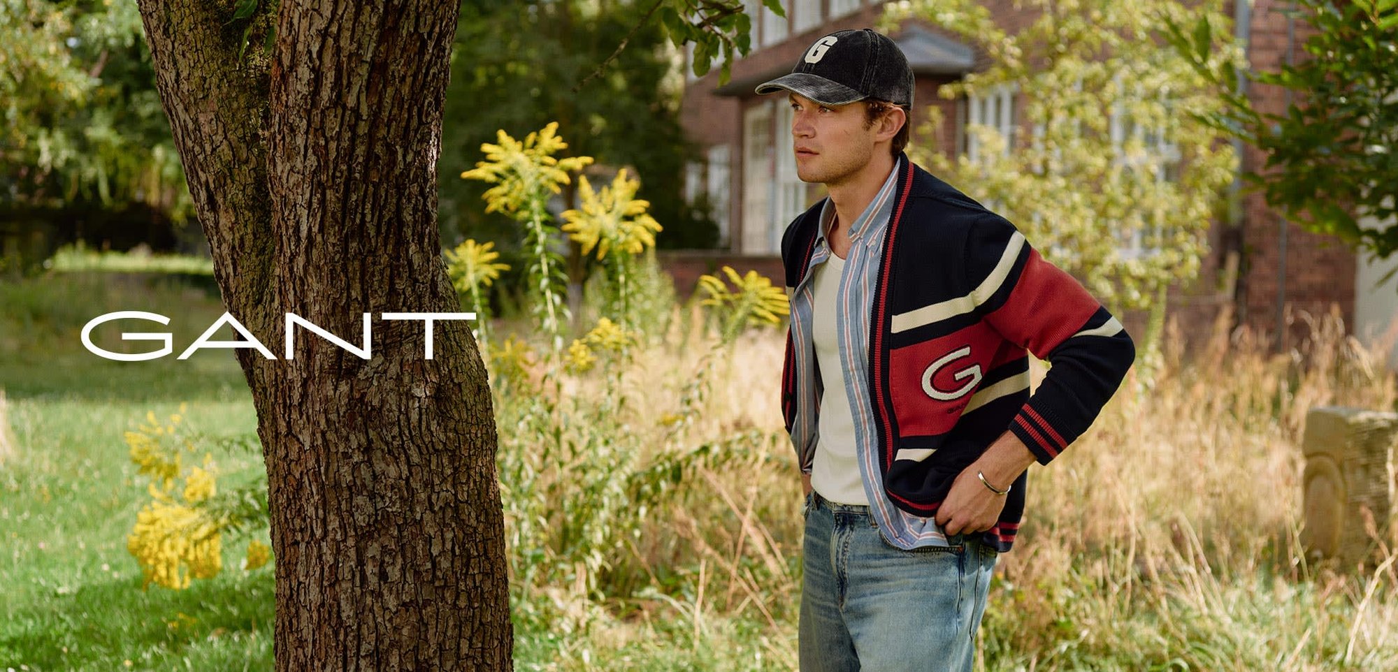 Navy, red, and cream striped cardigan over a white shirt; light blue jeans; black cap with a logo; standing beside a tree in grass.