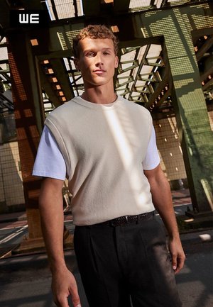 Beige gestricktes ärmelloses Oberteil über einem weißen T-Shirt, kombiniert mit dunkelgrauen maßgeschneiderten Hosen. Die Szene zeigt einstreuendes Sonnenlicht und eine industrielle Kulisse.