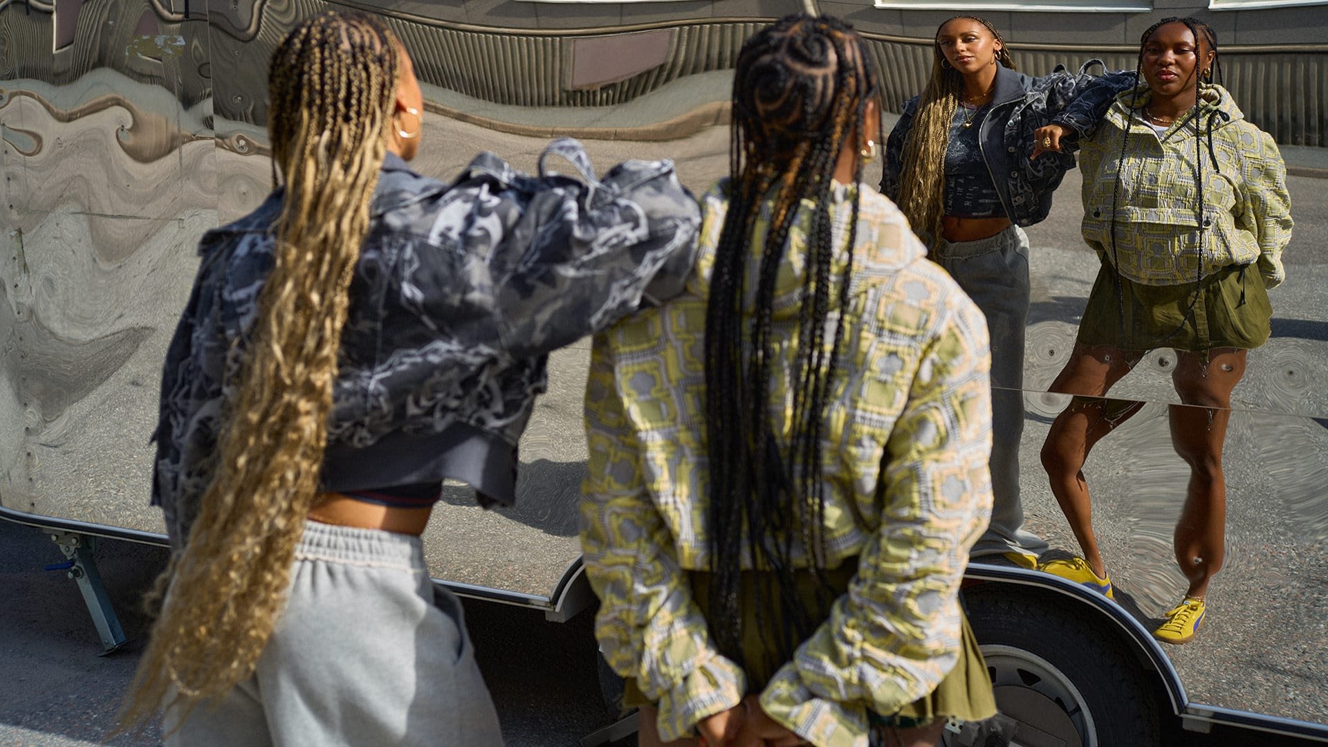 Twee personen in kortere jassen, de ene grijs met patronen en de andere geel met geometrische ontwerpen, poseren voor een spiegelend oppervlak.