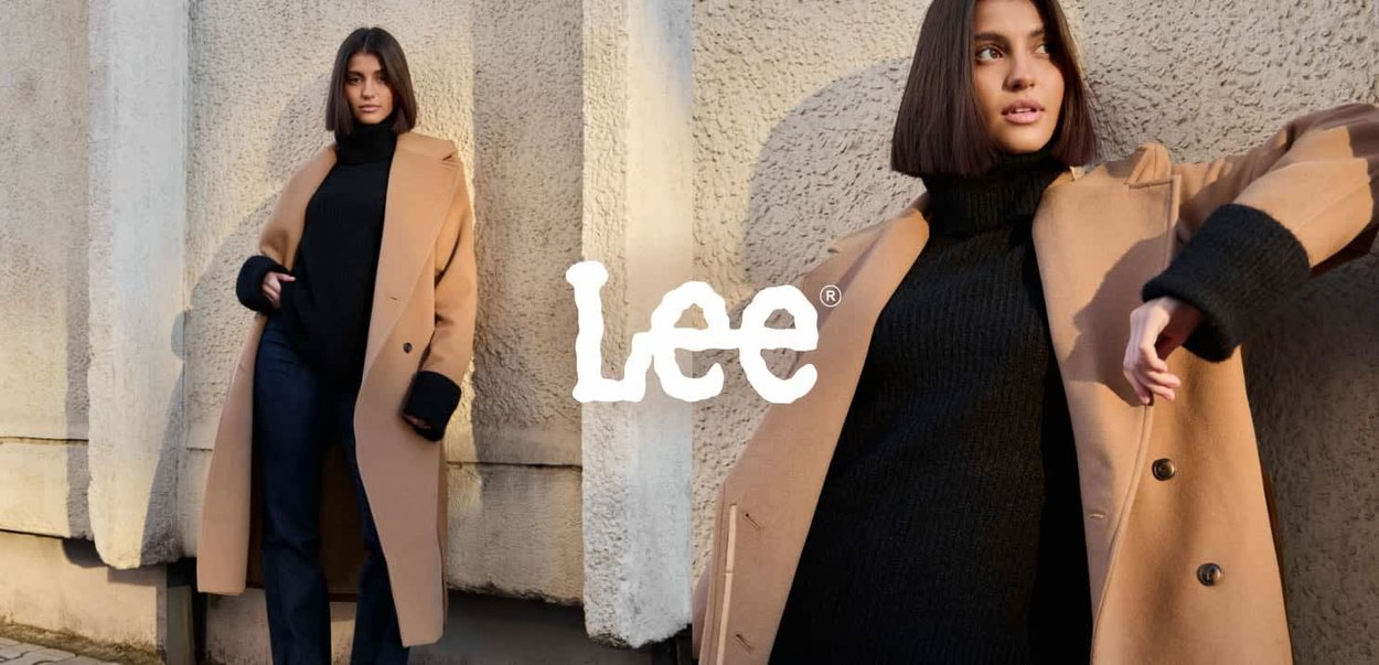 Abrigo largo de color marrón claro sobre un suéter de cuello alto negro, con puños anchos, botones y un corte holgado, combinado con vaqueros oscuros. El fondo es texturizado.