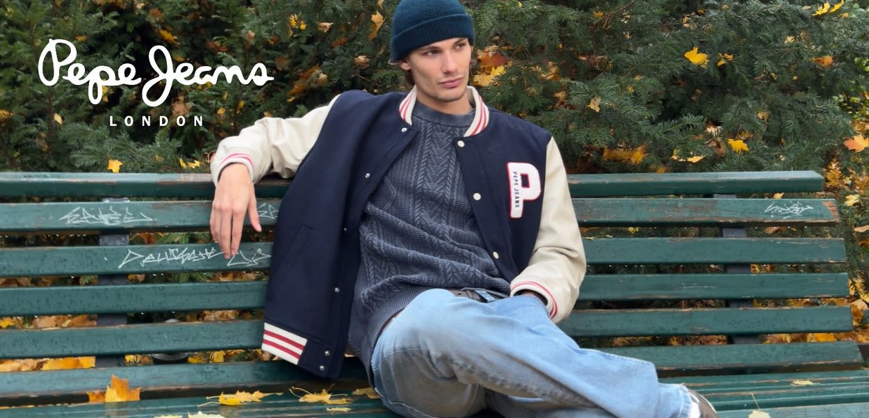Chaqueta varsity azul marino y crema con detalles en rojo sobre un suéter gris con estampado, combinada con jeans azul claro, sentado en un banco verde.