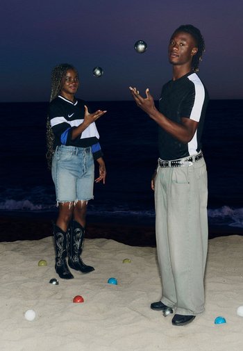 Dos personas en una playa de arena al atardecer, una con una camisa a rayas en blanco y negro y la otra con una camiseta negra, haciendo malabares con bolas metálicas.