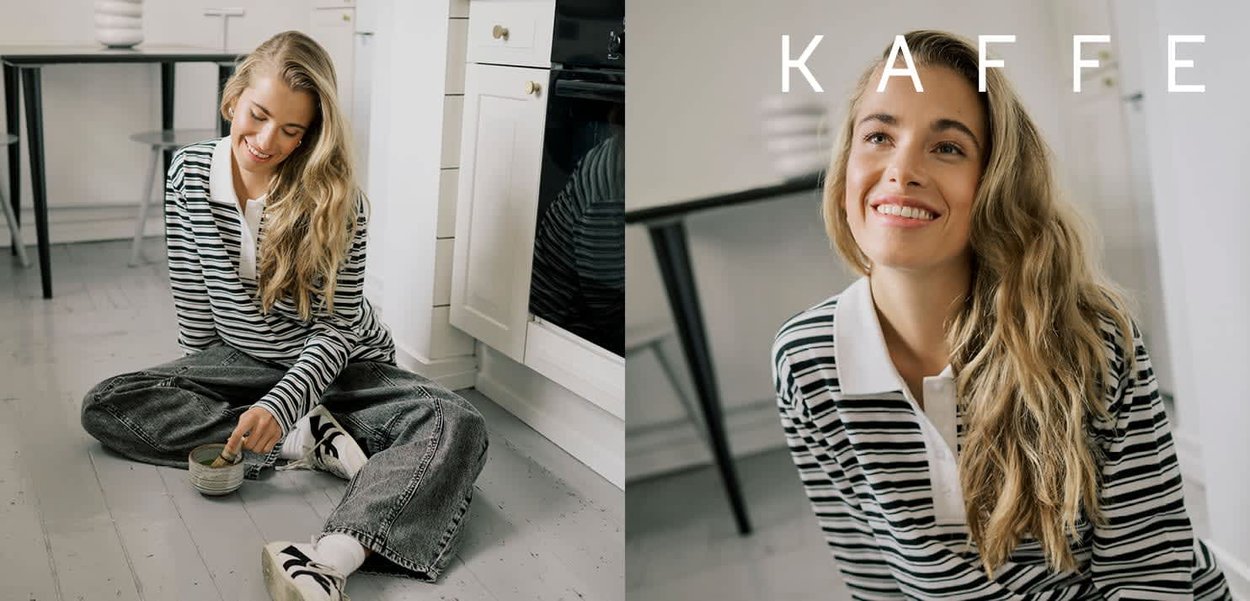 Junge Frau mit langen blonden Haaren und gestreiftem Hemd lächelt, sitzt mit überkreuzten Beinen auf einem Holzboden neben einem Küchenschrank und rührt in einer Tasse.