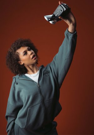 Sweatshirt zippé vert foncé avec un logo sur le devant, porté par-dessus un haut blanc. La personne tient un appareil photo au-dessus de sa tête devant un fond marron.