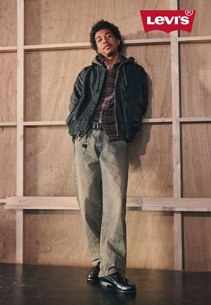 Junger Mann, der eine schwarze Jacke, kariertes Hemd, weite ausgewaschene Jeans und schwarze Schuhe trägt und vor einer Holzwand mit dem Levi's-Logo darüber steht.