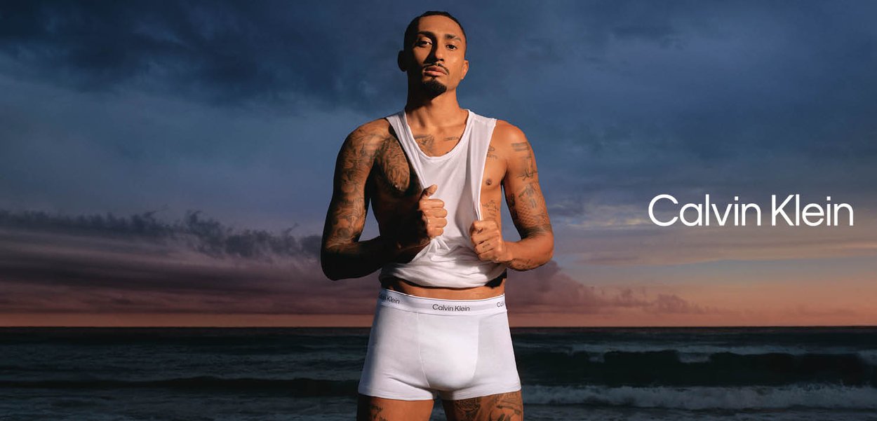 Mężczyzna z tatuażami podciąga biały tank top, ma na sobie białą bieliznę Calvin Klein o zachodzie słońca nad oceanem, logo Calvin Klein po prawej stronie.