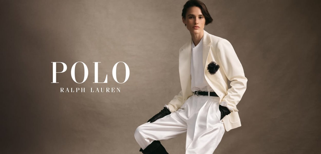 Valkoinen bleiseri mustalla kukkaakennolla, valkoinen paita, korkea vyötärö valkoiset housut ja mustat käsineet, neutraalia taustaa vasten.