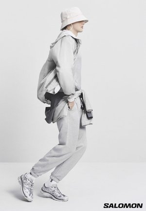 Person i lys grå afslappet påklædning, bøttehat og sølvfarvede sneakers, der går foran en ensfarvet hvid baggrund.