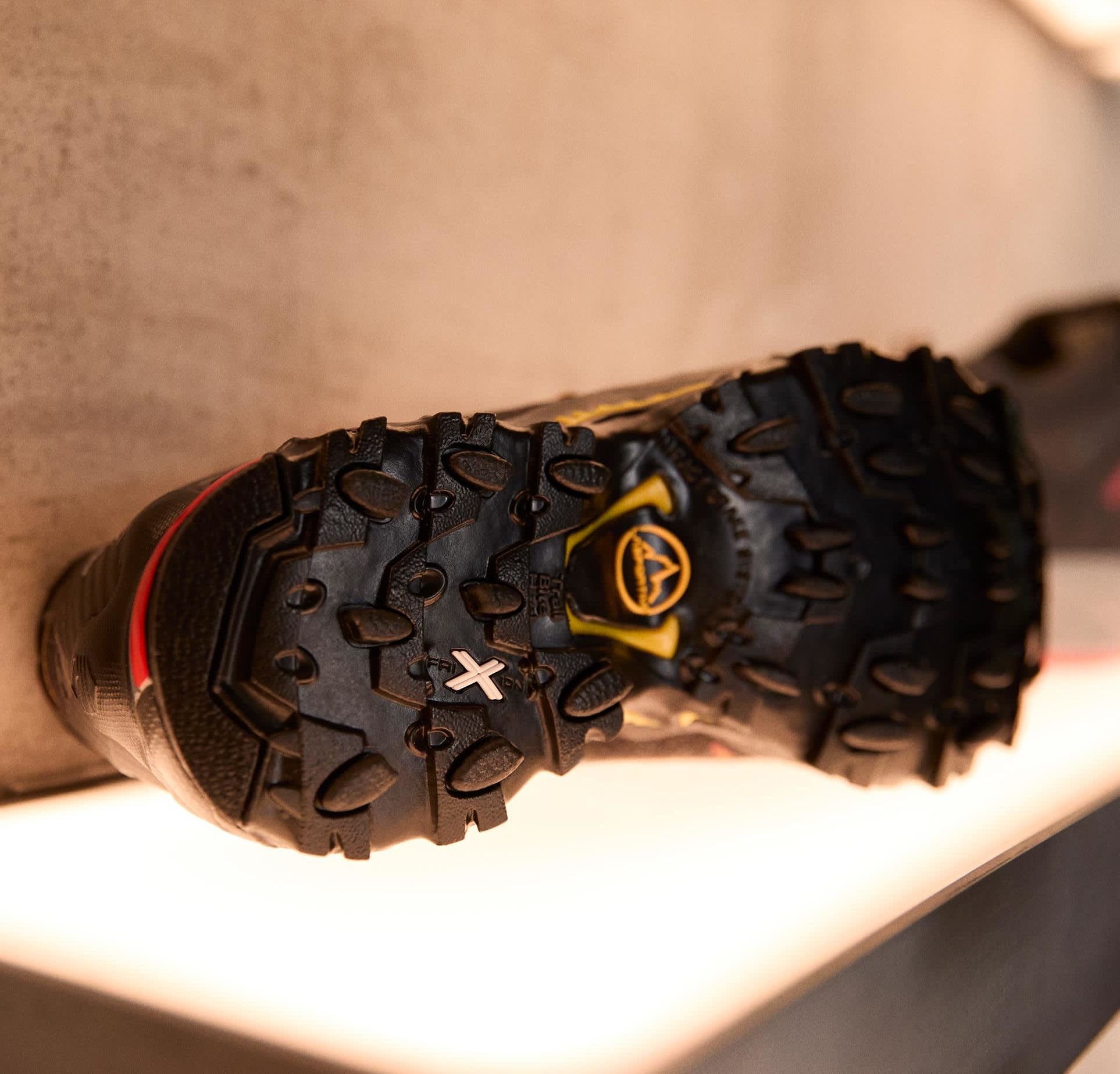 Schwarze Sportschuhsohle mit ausgeprägten Gummilaufflächen, gelben Akzenten und einer strukturierten Oberfläche. Besitzt ein 'X'-Markierung und Traktionsdetails.