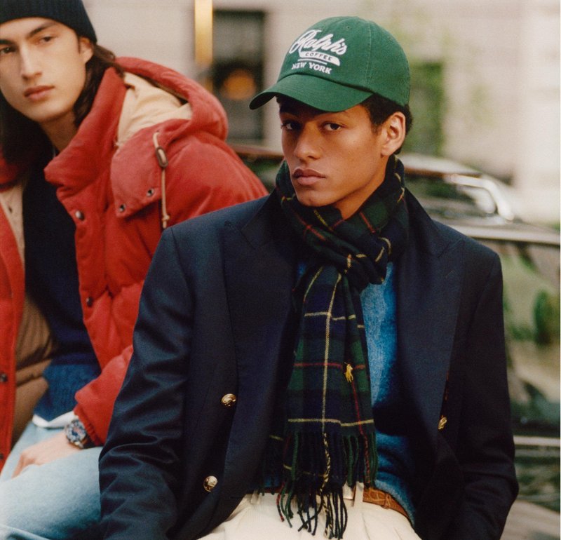 Two young men are seated outdoors. One wears a dark blazer, green plaid scarf, and green baseball cap; the other wears a red jacket.
