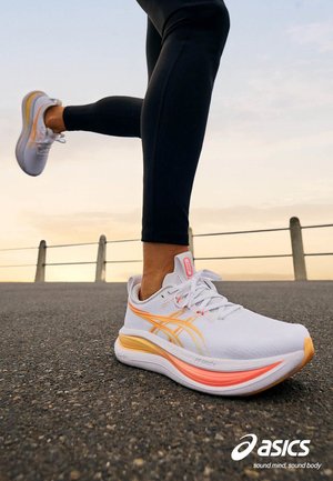 Corredor con zapatillas Asics blancas y naranjas y mallas negras en un camino asfaltado cerca de una barandilla al atardecer.