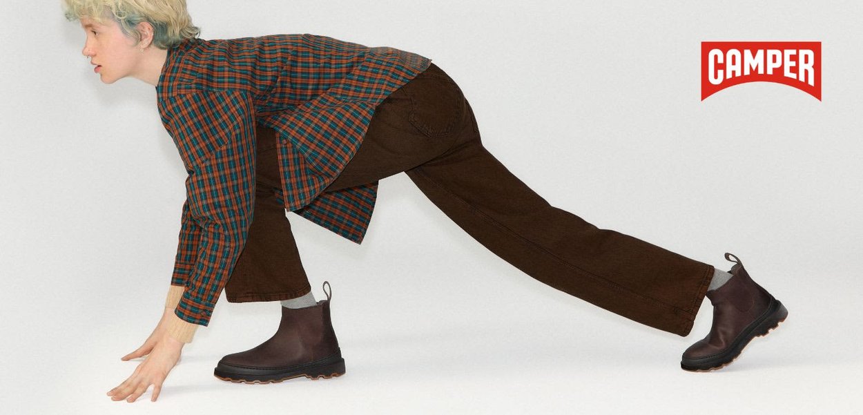 Person in karierter Bluse und brauner Hose, die sich nach vorne beugt, trägt dunkelbraune Stiefel mit groben schwarzen Sohlen, Camper-Logo in der Ecke.