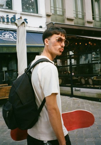 Zwarte rugzak op één schouder, wit T-shirt en rode skateboard onder de arm. Grote ronde zonnebril, stedelijke omgeving met tafels en stoelen.