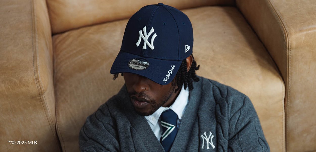 Uomo che indossa un berretto New York Yankees blu navy, maglione grigio con logo Yankees, camicia bianca e cravatta blu a motivi, seduto su un divano in pelle color cuoio.