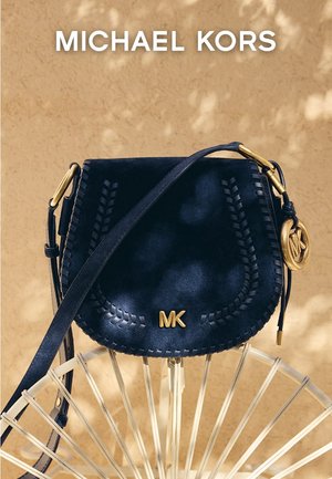 Schwarze Michael Kors Umhängetasche mit geflochtenem Besatz, goldfarbenem MK-Logo und goldfarbener Hardware, die an einem hellfarbenen Stuhl vor einem beigen Hintergrund hängt.