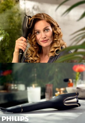 Donna con capelli ricci che utilizza un arricciacapelli automatico Philips, mostrato anche mentre è appoggiato su una superficie con oggetti da bagno sfocati sullo sfondo.