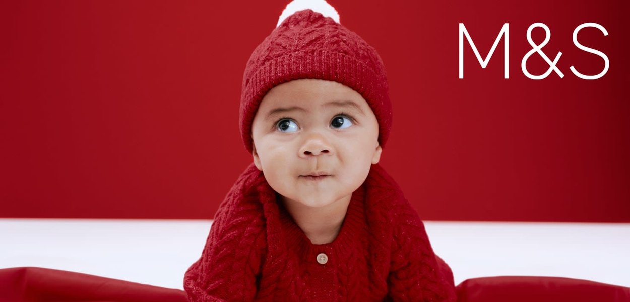 Czerwony sweter z dzianiny oraz dopasowana czapka z białym pomponem na czerwonym tle. Na swetrze wyróżnia się detal w postaci guzików.