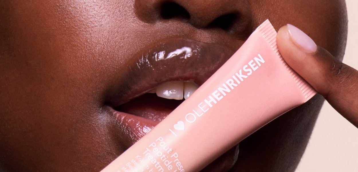 Un tube de traitement peptide préservant le pout d'Ole Henriksen de couleur pêche, tenu près des lèvres, lèvres brillantes visibles, texture de peau lisse en arrière-plan.