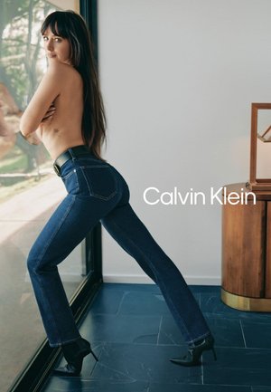 Mujer con cabello largo y oscuro, vestida con jeans azul oscuro y tacones negros, posando junto a una puerta de vidrio con el texto "Calvin Klein" visible.
