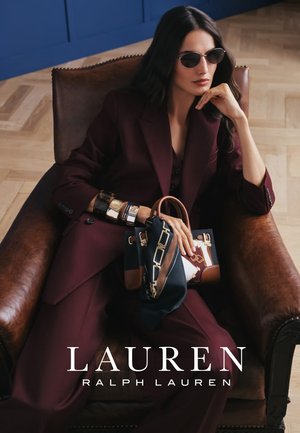 Mujer con traje color púrpura oscuro y gafas de sol sentada en un sillón de cuero, sosteniendo un bolso con estampado y pañuelos, con varias pulseras.