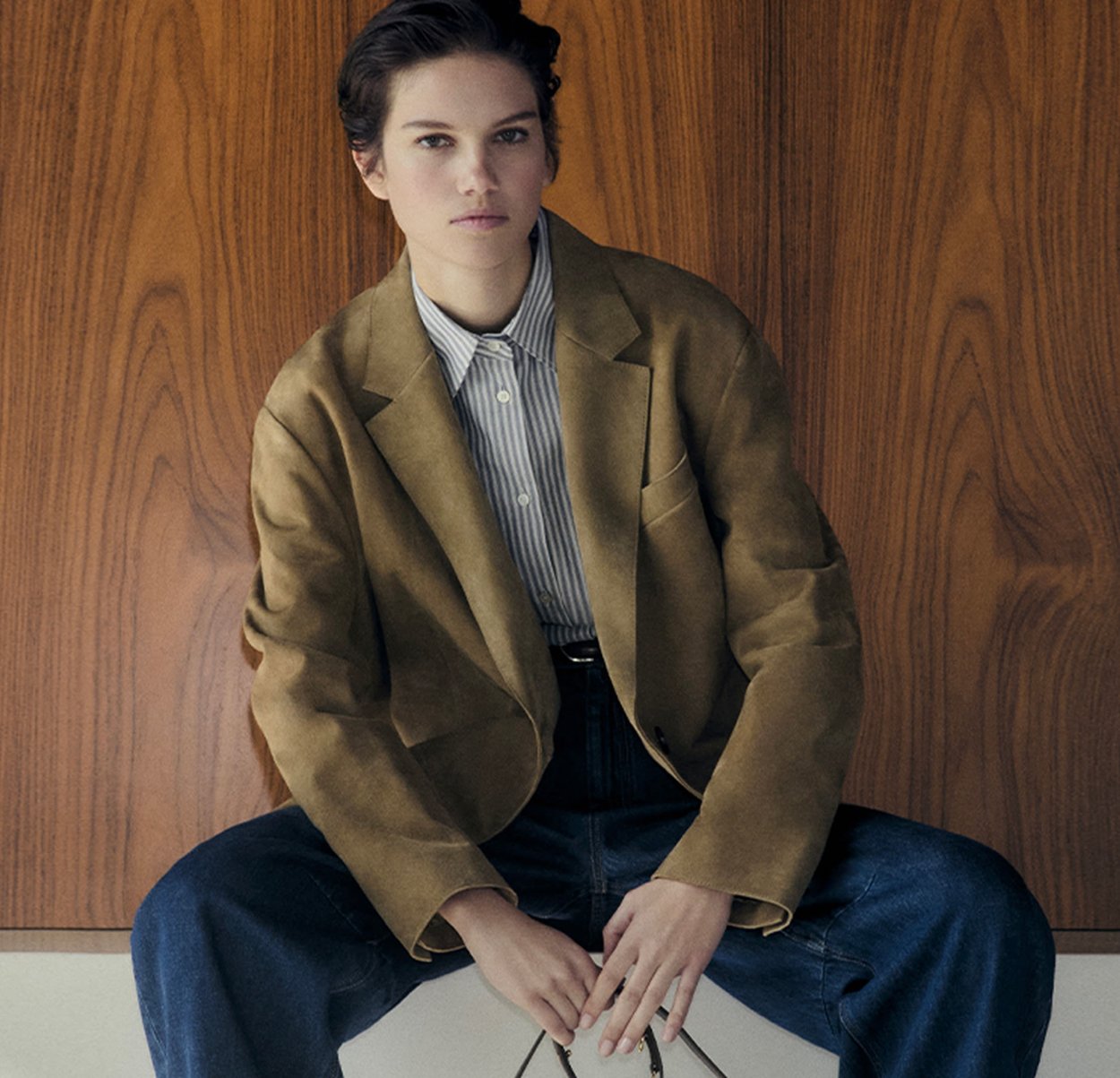 Olive green suede blazer over a striped shirt, paired with high-waisted blue jeans. The model sits on a white surface against a wooden backdrop.