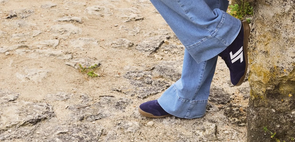 Chaussures en daim bleu marine avec un logo blanc, associées à un pantalon en denim clair à pattes d'éléphant, reposant sur une surface rocheuse et sablonneuse.