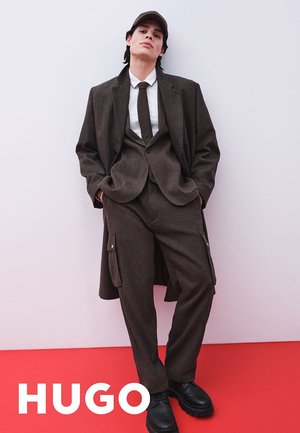 Traje marrón oscuro con abrigo oversize a juego, pantalones ajustados, chaleco abotonado, camisa blanca y zapatos negros frente a un fondo rosa.