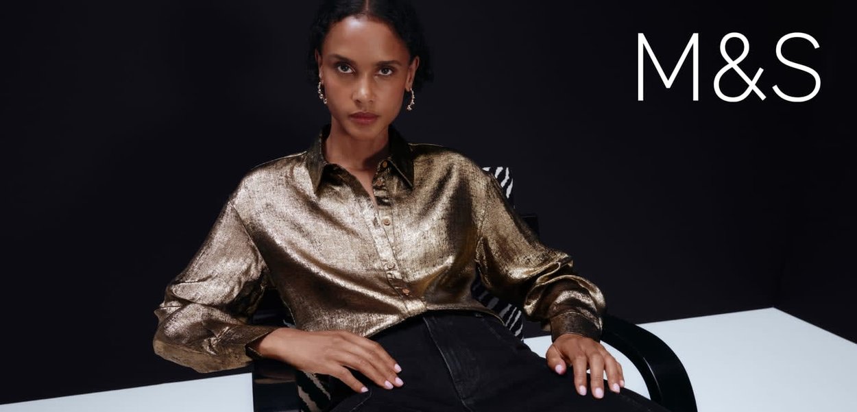 Chemise dorée métallique avec boutonnière avant, manches longues et col. Le modèle l'a assortie avec un jean taille haute foncé, assis sur une chaise.