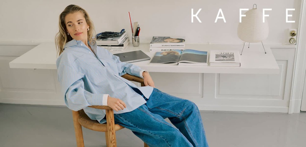 Hellblau gestreiftes Hemd, getragen mit locker sitzenden blauen Jeans aus Denim. Sitzt auf einem Holzstuhl neben einem minimalistisch gestalteten Schreibtisch mit Büchern und einer Lampe.