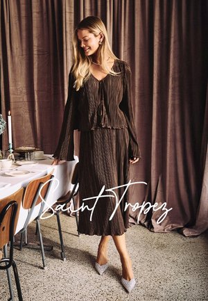 Braunes Faltenkleid mit gebundenem Ausschnitt und langen Ärmeln, kombiniert mit funkelnden spitzen Absätzen, vor einem sanft drapierten Hintergrund.