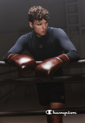 Pugile che indossa una maglia a maniche lunghe nera traspirante e guanti da boxe in pelle marrone, appoggiato a un ring con uno sfondo scuro.