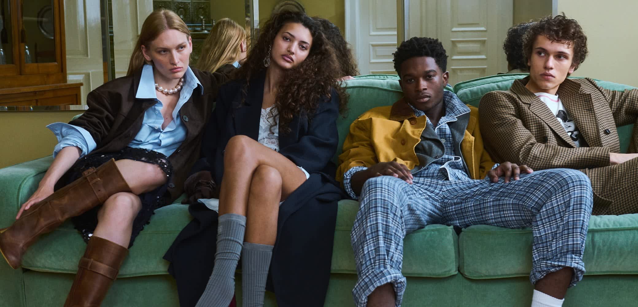 Fyra personer sitter på en grön soffa, klädda i olika mode stilar: läderstövlar, oversized ytterkläder, rutig och mönstrad textil, neutrala färger.