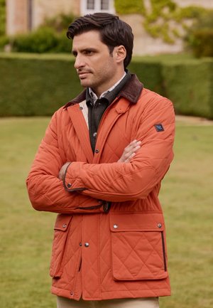 Chaqueta acolchada naranja con un cuello marrón, que presenta bolsillos frontales y cierre de cremallera. Llevada sobre un suéter gris con una camisa a cuadros.