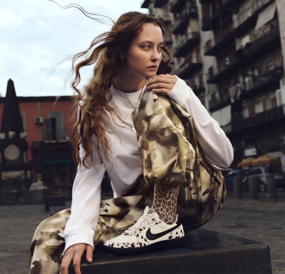 Sneakers Nike a pois bianchi e neri, pantaloni beige in camouflage e una maglietta a maniche lunghe bianca aderente. Capelli mossi, con uno sfondo testurizzato di edifici.