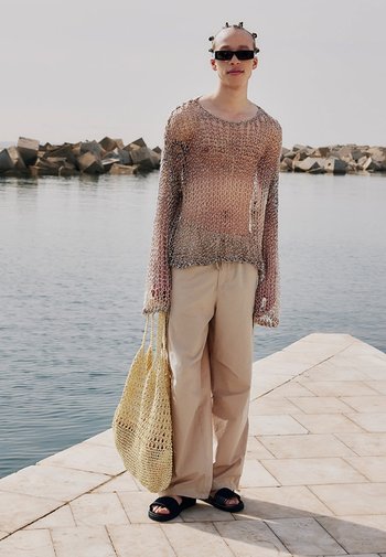 Pull en maille tricotée dans des nuances de marron, assorti à un pantalon beige à jambes larges. Tient un sac tissé jaune, porte des sandales noires à glissière.