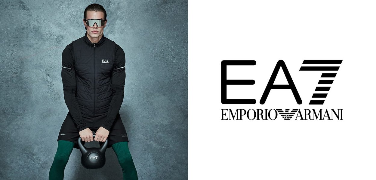 Gilet noir avec texture matelassée sur un haut à manches longues, associé à un short ajusté et des leggings verts, tenant un kettlebell, logo visible.