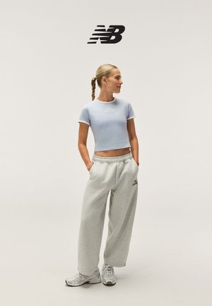 Donna in piedi con le mani nelle tasche, indossa una maglietta corta azzurro chiaro, pantaloni della tuta grigi, sneakers bianche e una coda di cavallo intrecciata, con logo New Balance sopra.