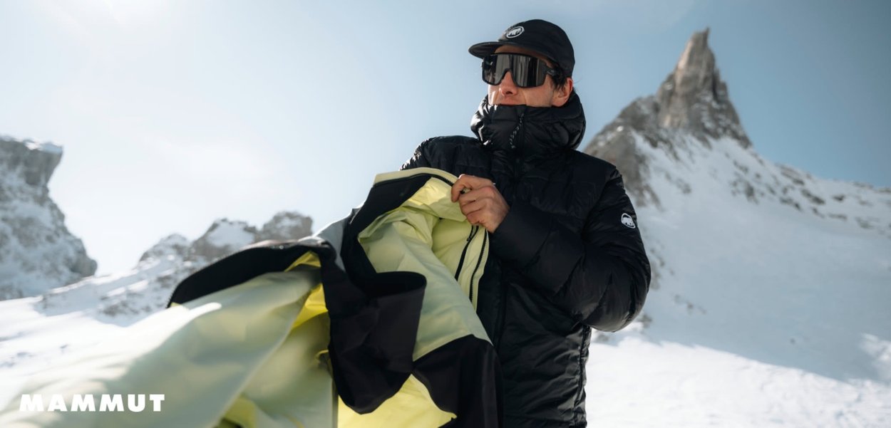 Czarna ocieplana kurtka z żółtą podszewką noszona przez osobę w czapce, trzymającą kurtkę w ośnieżonym krajobrazie z górskim szczytem.