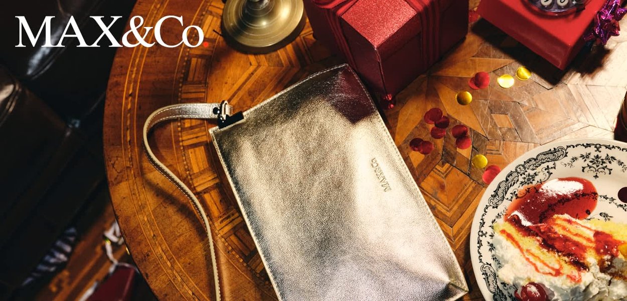 Borsa da polso color oro metallizzato su un tavolo di legno accanto a scatole regalo rosse, coriandoli sparsi e un piatto di dolce con crema e salsa rossa.