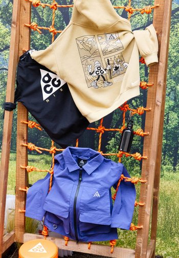 Sweat à capuche marron clair, t-shirt noir et veste zippée bleue avec logos Nike ACG affichés sur un cadre en corde orange en extérieur.