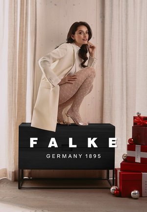 Pale beige coat over white top, model seated atop a black wooden box. She wears sheer, patterned tights and shiny silver heels. Red gift boxes nearby.