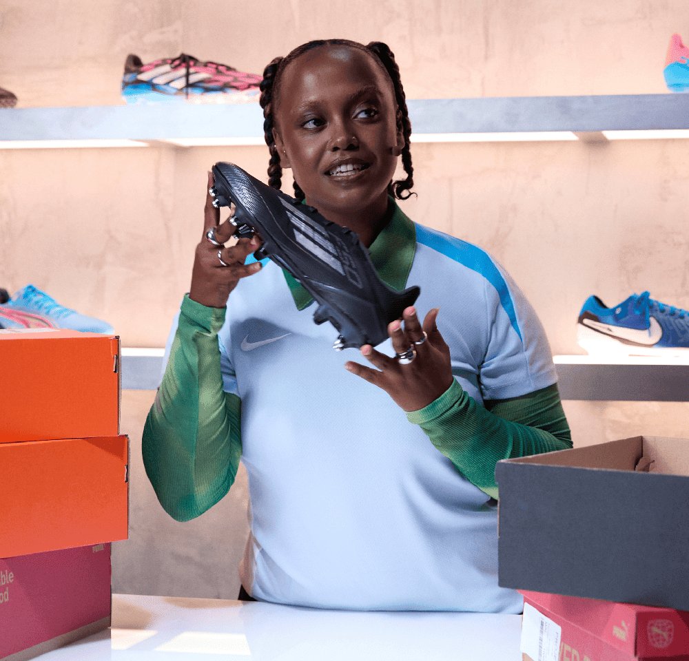 Schwarzer Fußballschuh, der in der Hand gehalten wird, mit einem eleganten Design. Drumherum sind orange und rote Kästchen. Im Hintergrund sind weitere farbenfrohe Sportschuhe zu sehen.