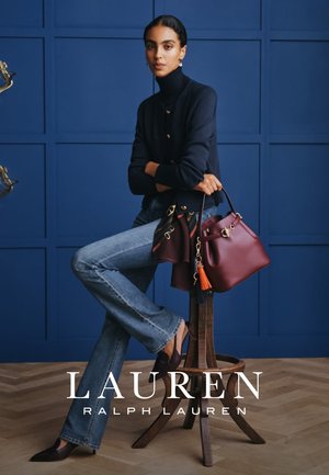 Donkerrode leren handtas met een gouden accent en een oranje kwastje, gecombineerd met uitlopende spijkerbroeken en een donker vest tegen een blauwe achtergrond.