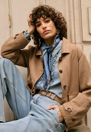 Junge Frau mit lockigem Haar, die eine hellbraune Jacke, ein blaues Denim-Outfit und einen blau gemusterten Schal trägt und entspannt an einer beigen Wand sitzt.