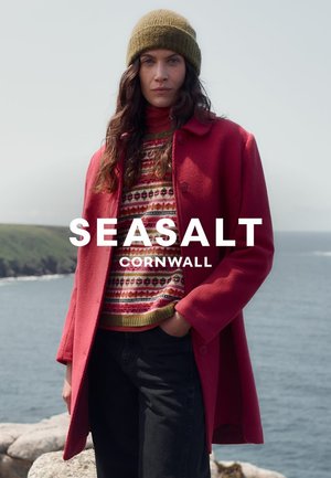 Manteau en laine rouge avec de gros boutons, porté sur un pull en tricot à motifs. Le modèle porte un bonnet vert olive. L'océan est visible en arrière-plan.