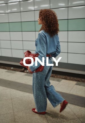 Donna con capelli ricci che indossa un completo di jeans a motivi e scarpe rosse, con una borsa rossa in mano, che cammina in una stazione della metropolitana piastrellata.