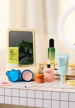 Verschiedene Hautpflegeprodukte, darunter ein Sonnenschutz-Stick, Serum, Feuchtigkeitscreme und Gesichtscreme, auf einer gefliesten Oberfläche zusammen mit einem Spiegel und Büchern arrangiert.