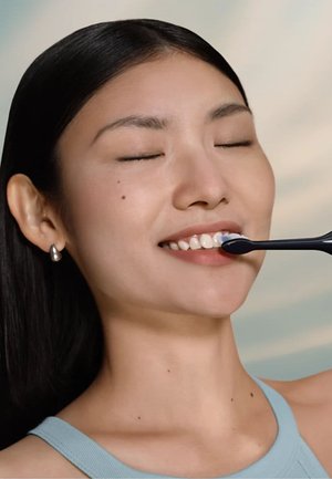 Brosse à dents électrique avec un manche noir et des poils bleus, tenue près d'une bouche souriante. L'arrière-plan présente des couleurs pastel douces et floues.